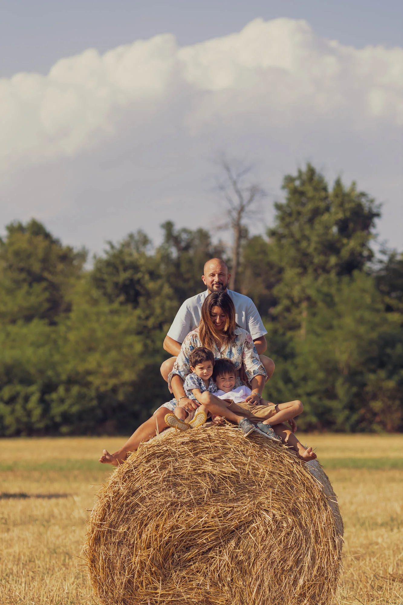 Famiglia seduta in cima a una grande balla di fieno, sotto un cielo con nuvole bianche